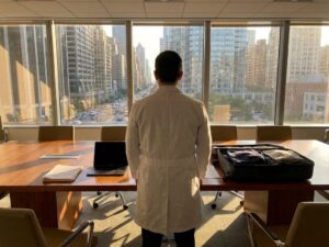 Researcher in lab coat at conference room window overlooking city, with laptop and packed suitcase on table behind them.